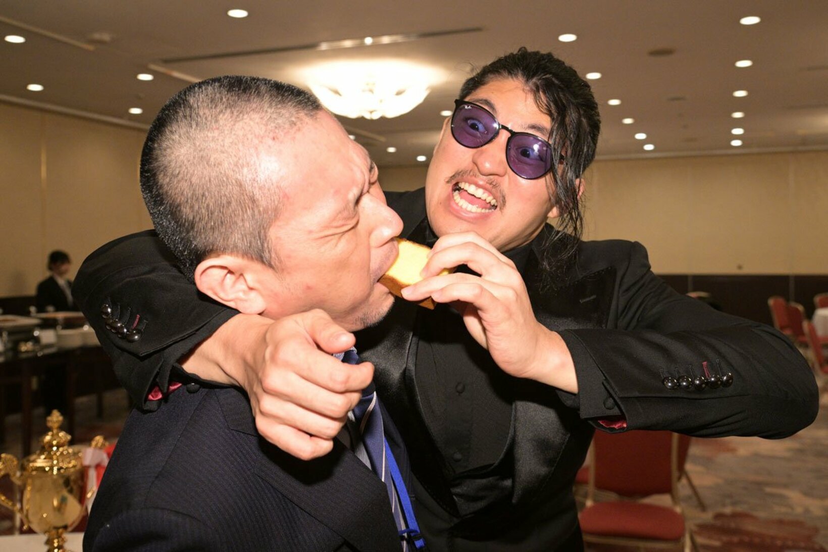 記者の口にカステラを押し込むOZAWA（右）