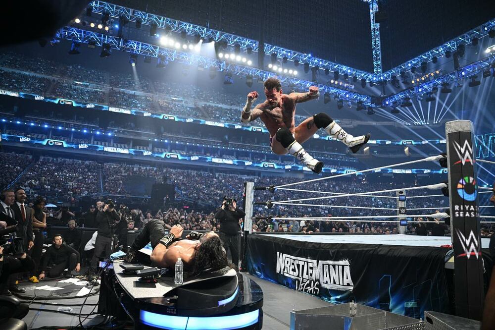 実況席上のローマン・レインズ（下）にCMパンクはエルボードロップ（©WWE）