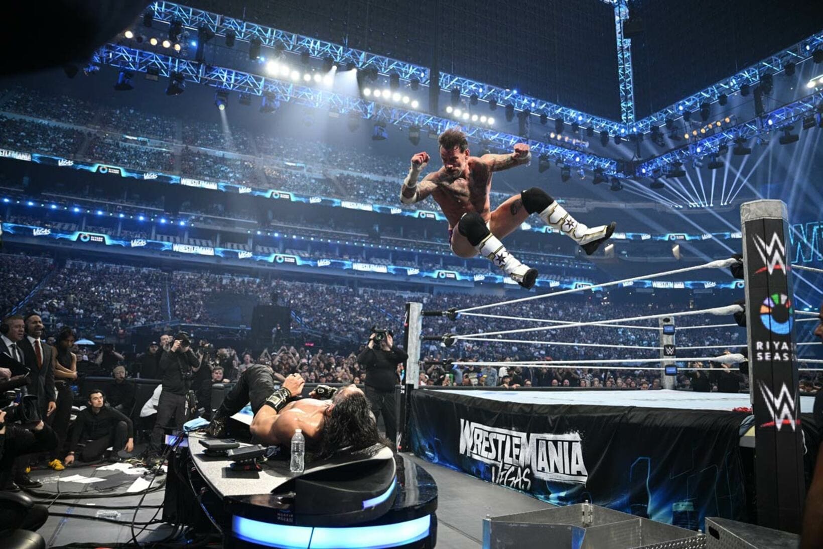 実況席上のローマン・レインズ（下）にCMパンクはエルボードロップ（©WWE）